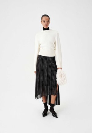 Mujer con suéter blanco sobre un cuello alto negro, falda negra de lunares con abertura lateral, sosteniendo un bolso de mano blanco y esponjoso, tacones negros con calcetines.