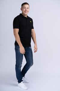 Čierna polo košeľa so žltým logom, v kombinácii s modrými džínsami a bielymi teniskami. Strih je moderný a látka vyzerá hladko.