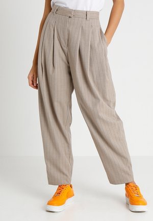 Beżowe spodnie w paski z wysokim stanem, plisowanym przodem i bocznymi kieszeniami, zestawione z jaskrawymi pomarańczowymi trampkami.