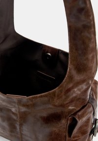 Bruine leren schoudertas met een gladde textuur, grote opening, drukknoopsluiting en een klein binnenvak. Beschikt over een zijvak en metalen hardware.