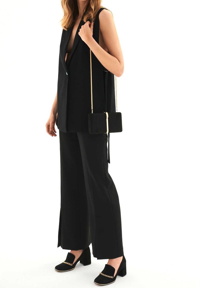 Femme portant un tailleur noir sans manches avec un pantalon à jambes larges, tenant un petit sac noir avec chaîne dorée et portant des chaussures à talons blocs noires avec détail chaîne dorée.