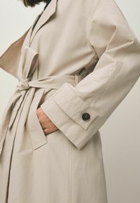 Beige trenchcoat med bælte, med en afslappet pasform, lange ærmer og en enkelt knap ved manchetterne. Glat, letvægtsstof.