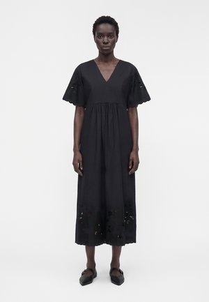 Donna in piedi di fronte che indossa un vestito midi nero con scollo a V, maniche corte svasate e dettagli a fiori traforati sull’orlo e sulle maniche.