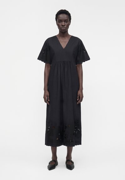 Femme debout face à l’avant, portant une robe midi noire à col en V, manches courtes évasées et détails découpés floraux à l’ourlet et aux manches.