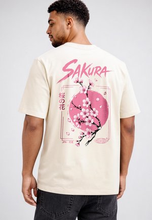 Man wearing cream t-shirt with pink cherry blossom branch and large circle design and "SAKURA" text on the back.