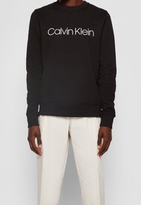 Sudadera negra de cuello redondo, hecha de una mezcla de algodón, con el logo blanco de "Calvin Klein" en la parte frontal y puños y dobladillo acanalados.