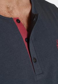 Dark grey T-shirt with a red accent at the collar, featuring a three-button placket and embroidered details. Smooth cotton material.