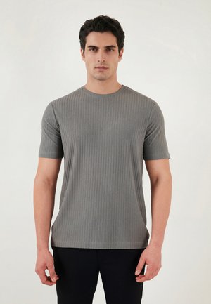 Jeune homme aux cheveux foncés portant un t-shirt gris côtelé à manches courtes et un pantalon noir, debout devant un fond clair uni.