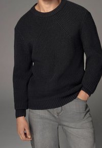 Modèle portant un pull noir texturé et un jean gris clair, debout contre un fond neutre, avec une main posée dans la poche du pantalon.