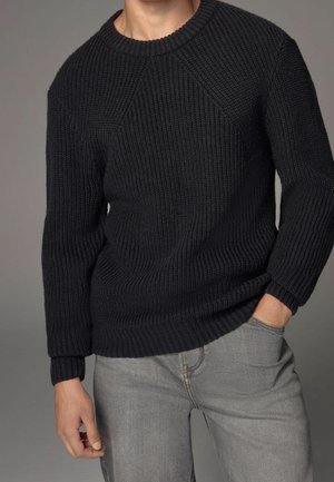 Modèle portant un pull noir texturé et un jean gris clair, debout contre un fond neutre, avec une main posée dans la poche du pantalon.
