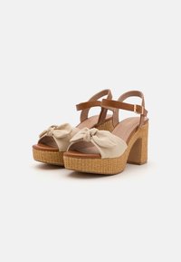 Beige platform sandals with a knotted fabric upper, brown adjustable ankle strap, and a woven texture on the block heel.