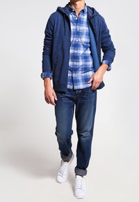 Marinblå zip-hoodie över en blå rutad skjorta, matchad med mörkblå jeans och vita sneakers.