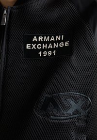 Giacca in mesh nera con etichetta "ARMANI EXCHANGE 1991" e logo "AX" testurizzato. Chiusura con zip sulla parte anteriore.