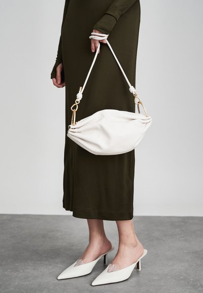 Baltas ādas rokassomi ar noapaļotu, krokotu dizainu, ko tur knašķis plecu siksna ar zelta aksesuāriem.