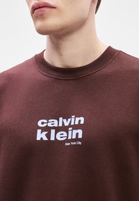 Καφέ φούτερ με κοπή crew και λευκό κείμενο "Calvin Klein New York City". Το ύφασμα φαίνεται μαλακό με λεία υφή.