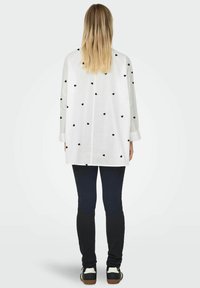 Camicia bianca a maniche lunghe con motivi a cuori neri. Indossata con leggings scuri e sneakers, vista da dietro. Tessuto morbido e vestibilità rilassata.