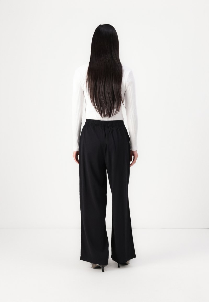 Schwarze weit geschnittene Hosen mit einem elastischen Bund, kombiniert mit einem enganliegenden weißen Langarmoberteil, von hinten gesehen. Glatte Textur.