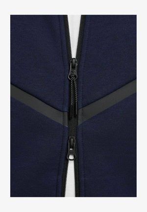 Marineblå hættetrøje med lynlås og sorte lynlåse og detaljer, der har en blød stoftekstur og et kontrasterende hvidt indre lag.