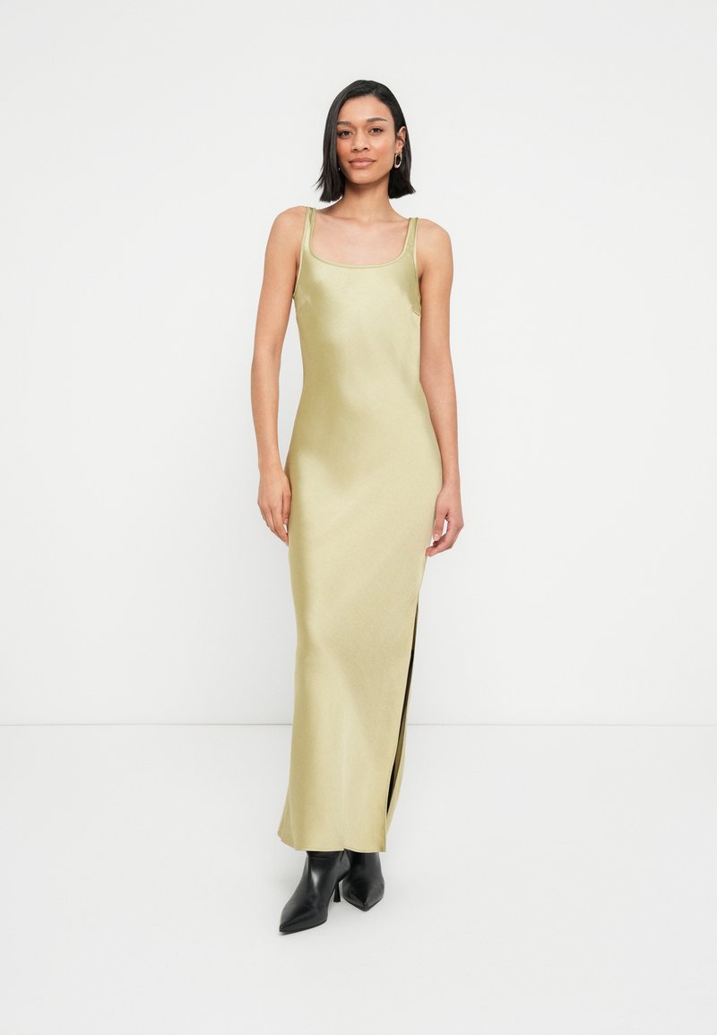 Μακρύ φόρεμα από σατέν σε ελιά πράσινο με τετράγωνη λαιμόκοψη, λεπτές τιράντες, σκίσιμο στο πλάι και λεία υφή. Συνδυάζεται με μαύρα μυτερά μποτάκια αστραγάλου.