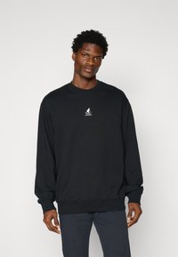 Melns džemperis, kas izgatavots no mīkstas auduma; tam ir apaļa kakla izgriezuma, garas piedurknes un neliels balts logo krūtīs. Gluda tekstūra.