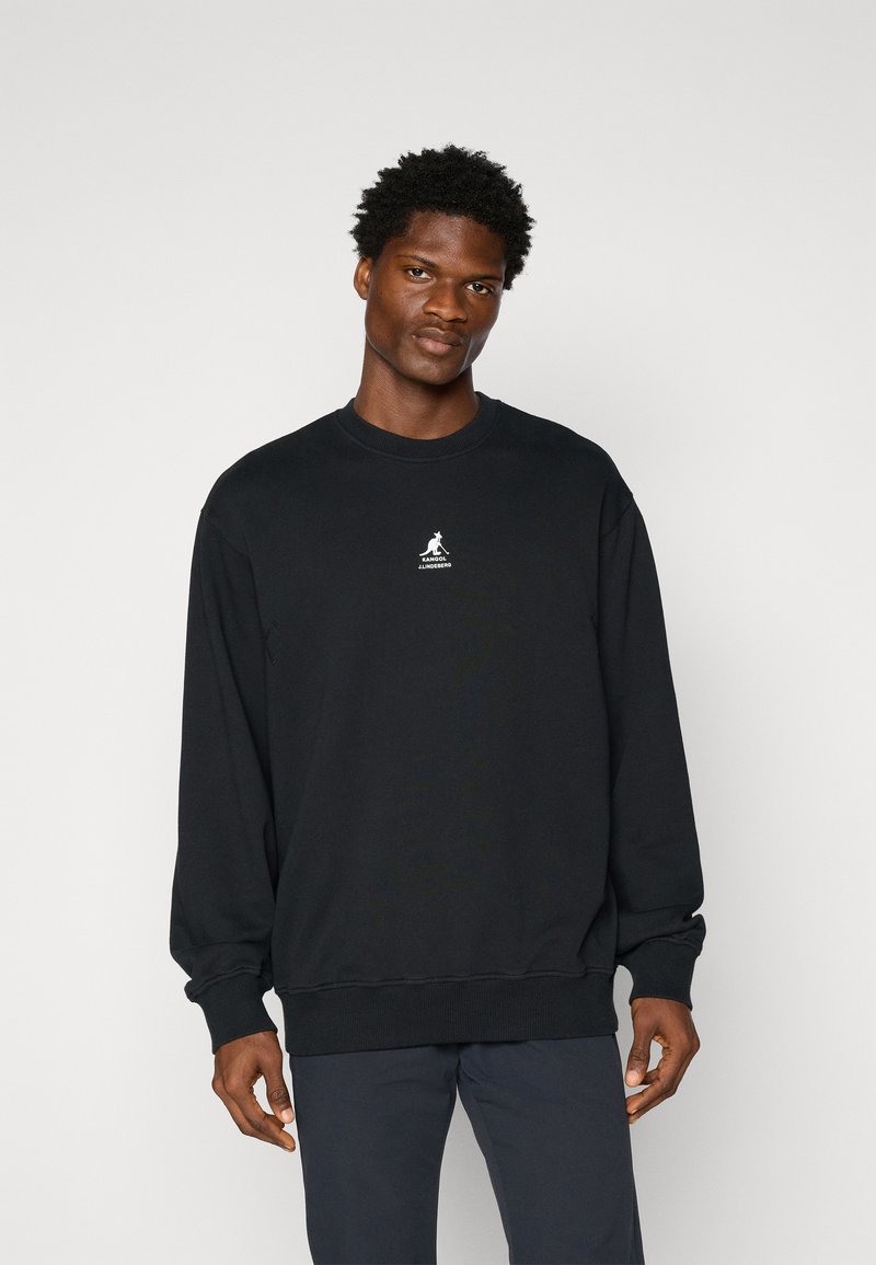 Melns džemperis, kas izgatavots no mīkstas auduma; tam ir apaļa kakla izgriezuma, garas piedurknes un neliels balts logo krūtīs. Gluda tekstūra.