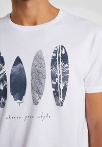 Weißes Baumwoll-T-Shirt mit blauen Grafiken von Surfboards, die verschiedene Muster aufweisen, und dem Text "Wähle deinen Stil" am unteren Rand.