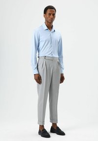 Chemise bleu clair à manches longues avec fermeture à boutons, associée à un pantalon plissé gris clair avec boutons latéraux et revers pliés. Mocassins noirs.