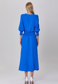 Robe bleue à manches longues avec une taille smockée, en tissu fluide et de longueur midi. Portée avec des sandales blanches à talons. Texture lisse.