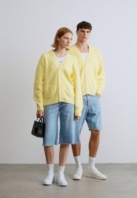 Κίτρινα ριγέ καζάκια με κουμπιά, φορεμένα πάνω από λευκές μπλούζες, σε συνδυασμό με τζιν σορτς. Αξεσουάρ περιλαμβάνουν μια μικρή μαύρη τσάντα χειρός.