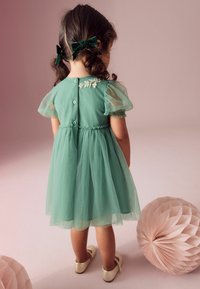 Mäta zelené tylové šaty s balónovými rukávmi, zapínaním na gombíky a kvetinovými ozdobami na zadnej strane. Krémové baletky dopĺňajú outfit.
