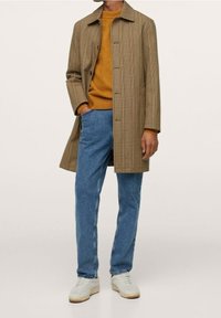 Beżowy płaszcz w kratę o prostym kroju, noszony na musztardowym swetrze, połączony z niebieskimi dżinsami i białymi trampkami.