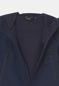 Navy blue softshell jacket with fleece lining, two vertical zippered chest pockets, and branded tag inside the collar.