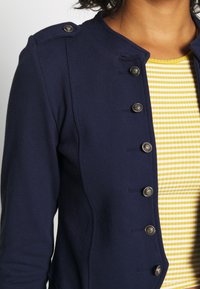 Gros plan d'une personne portant une veste bleu marine avec des boutons argentés par-dessus une chemise à rayures jaunes et blanches, montrant le haut du torse et le cou.