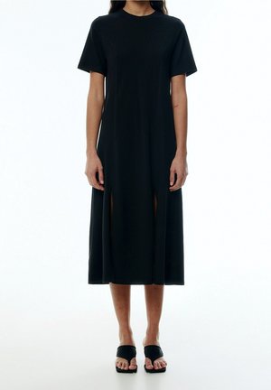 Donna che indossa un abito midi nero a maniche corte con spacchi frontali, abbinato a sandali neri con tacco quadrato e punta aperta, in piedi davanti a uno sfondo bianco.