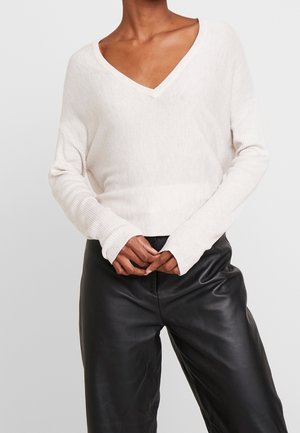 Helle beige V-Ausschnitt-Strickjacke mit gerippter Textur und langen Ärmeln, kombiniert mit schwarzen Lederhosen, die eine lockere Passform zeigen.