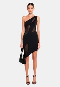 Abito nero a spalla unica con pannelli trasparenti, orlo asimmetrico e texture liscia. Modella che tiene una piccola borsa nera con finitura lucente.