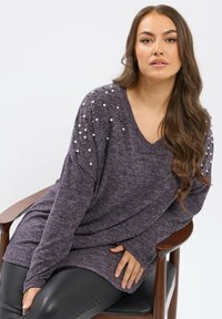 Maglia lunga viola con scollo a V e spalle decorate, che presenta dettagli in perle e accenti metallici, che scende morbida su un pantalone nero simile alla pelle.