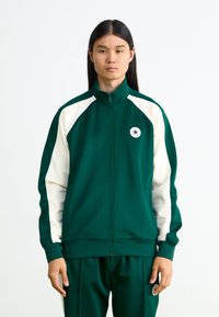 Πράσινη ζακέτα με φερμουάρ, λευκά ράγκλαν μανίκια, στρογγυλό λογότυπο με έναν μπλε αστέρα και υφή υφάσματος. Συνδυασμένη με αντίστοιχες πράσινες παντελόνες.