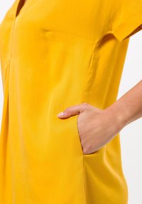 Gelbes Kleid aus leichtem Stoff, mit einem schlichten Design, kurzen Ärmeln, einer Tasche an der Seite und einer lockeren Passform.