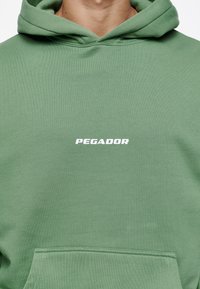 Grøn hættetrøje med en kængurulomme, tekstureret stof og en hvid "PEGADOR"-logo på brystet. Dobbeltsiddet hætte for ekstra varme.