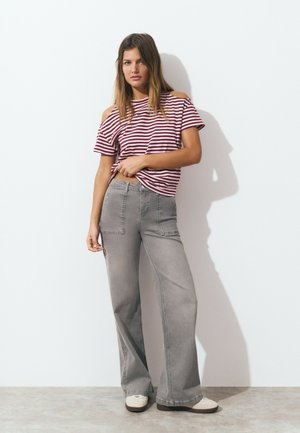 Femme portant un haut à rayures bordeaux et blanches, un jean large gris et des baskets crème, se tenant contre un mur blanc.