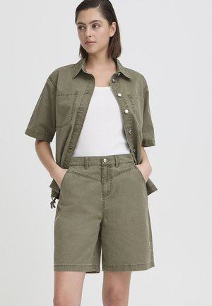 Kvinde iført olivengrøn kortærmet skjorte med knapper og matchende shorts til knæet, kombineret med en hvid ærmeløs top, stående med hænderne i lommerne.