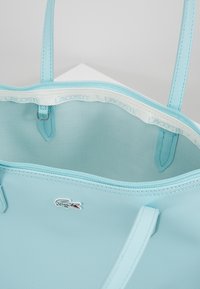 Jasnoniebieska torba typu tote wykonana z materiału teksturowego, z białym wnętrzem ozdobionym logo "LACOSTE", górnym zamkiem błyskawicznym oraz detalem logo.