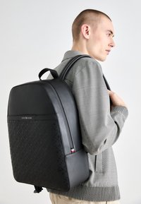 Sac à dos noir avec un design texturé, des accents en cuir lisse et des bretelles rembourrées réglables. Logo affiché au centre en haut.