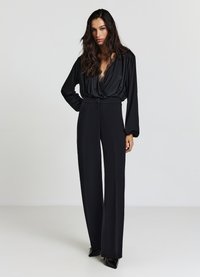 Schwarzer Jumpsuit mit tiefem V-Ausschnitt, langen Ärmeln und weitem Bein. Hergestellt aus glattem Stoff mit zarten Spitzenbesätzen.