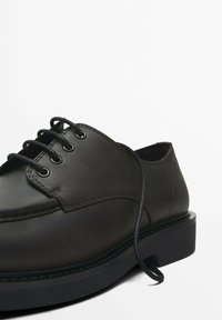 Black leather shoe with round toe, six eyelets, and black laces. Features a pronounced stitched welt and thick rubber sole.