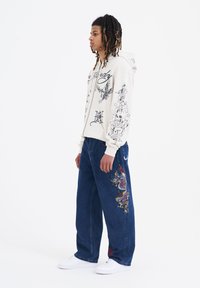 Sudadera crema con estampados gráficos, combinada con pantalones de denim azul oscuro que presentan un bordado colorido de dragón. Acompañada de zapatillas blancas.