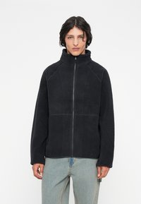 Giovane che indossa una giacca in pile nera con cerniera e colletto alto, e jeans azzurri chiari, in piedi davanti a uno sfondo bianco uniforme.