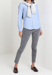 Camicia button-up blu con un motivo sottile, maglione grigio appoggiato intorno al collo, pantaloni slim-fit grigi e mocassini bordeaux.