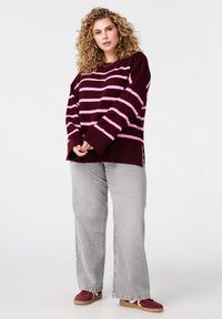 Gebreide trui met bordeauxrode en roze strepen, oversized pasvorm, met lange mouwen en zijsplitten, gecombineerd met lichtgrijze jeans met wijde pijpen.